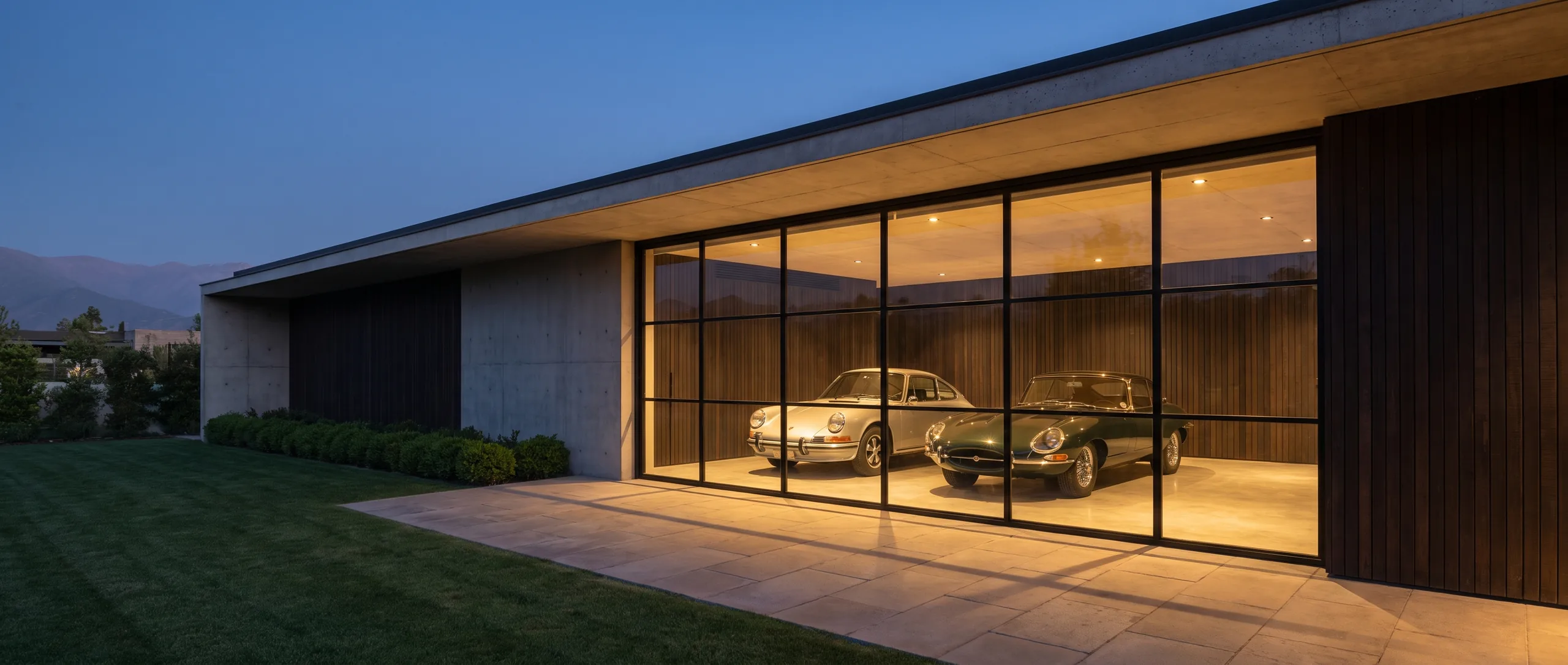 Garaje de colección al atardecer: volumen de hormigón y madera con un gran vano vidriado iluminado, dos autos clásicos adentro